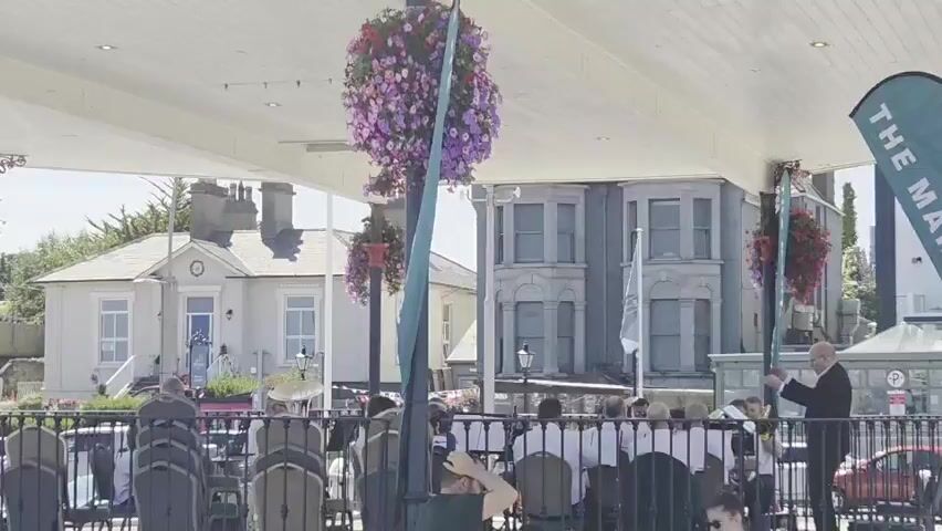 A group of people are gathered outdoors under a white canopy, with hanging baskets of purple and pink flowers. In the foreground, chairs are set up. A man in a dark suit stands on the right, facing away from the camera, and appears to be directing the group. The background shows buildings, including a light-colored house with columns and a grey, ornate building. A green banner with the words "THE MART" is visible on the right. The scene appears to be a performance or a formal gathering.