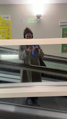 The video shows a woman riding an escalator in what appears to be a store. She is reflected in a glass panel. She is wearing a long, olive-green coat, a pink hoodie, jeans, and studded shoes. She has headphones on. She is holding a bottle of blue cleaning product in one arm and a phone in the other, taking a selfie. The escalator is going up. There is a green poster and a sign that reads "CREDITO COOPERATIVO" on the wall behind her.