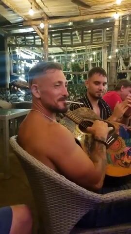 Here is a description of the video:

The video shows four men sitting around a table on a covered patio. The man closest to the camera is shirtless and sitting in a wicker chair, playing a colorful acoustic guitar. He has short hair with a design shaved into the side, and is wearing a silver chain necklace and a beaded bracelet. A cigarette is placed behind his ear. The other three men are sitting further away and are dressed in casual shirts. They are clapping and singing along to the music. The patio has a wooden frame and a bamboo roof, with string lights hanging around. A ceiling fan is turning overhead.