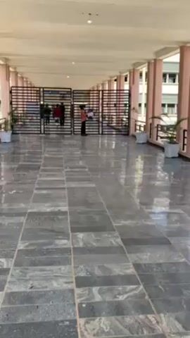 The video shows a long, covered walkway with a tiled floor. The walkway leads to a gate with several people standing near it. The area is well-lit. The building on the right has windows. Planter pots are placed along the side of the walkway. A person wearing a red shirt and a light blue hat decorated with colorful balls walks toward the gate.