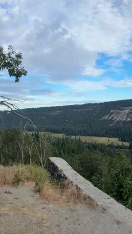 A landscape video shows a valley filled with evergreen trees. A road winds through the valley, and a small lake or pond is visible. The sky is blue with scattered clouds. In the foreground, there is a low stone wall and some dry grass.