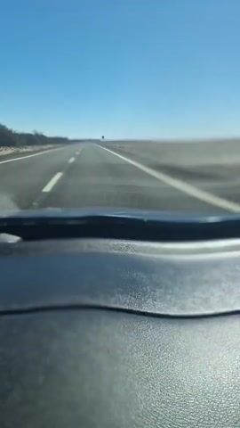 The video shows a first-person view from inside a vehicle, looking out through the windshield. The vehicle is driving on a paved road with white lane markings. The road stretches into the distance under a clear blue sky. The landscape on either side of the road appears to be flat and open, with some sparse vegetation and what look like utility poles or windmills in the distance. The dashboard of the vehicle is visible in the foreground, in focus and textured. The overall impression is of a long drive on an open highway.