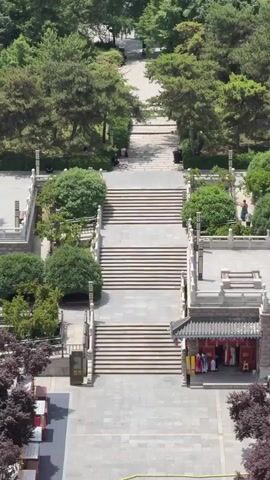 The camera is set high and pointed down a wide set of stone steps towards a long straight road lined with trees. At the base of the stairs, a building with a traditional Chinese style roof and red accents is visible.  To the right and left of the steps are landscaped areas with bushes and small trees. In the distance, cars travel along the road which is flanked by more trees and buildings. A few people are visible walking in the park area at the top of the steps.