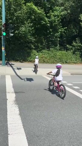 This media file captures a sunny day scene in Port Moody, Canada, featuring a group of people, primarily children, cycling on a paved road with a pedestrian crossing. The sunlight creates distinct shadows, suggesting it is midday. The background is dominated by lush green trees, indicating a natural or park-like setting.

The main subjects are several individuals on bicycles. Two young girls are prominently featured, one wearing a pink helmet and a light blue outfit, the other in a white shirt and pink helmet. They are riding their bikes through a crosswalk. Behind them, two adults are also on bicycles, one wearing a blue backpack and the other a grey backpack. Further in the background, another child on a bicycle is visible.

The activity is clearly cycling, with the group navigating a road and a pedestrian crossing. The presence of traffic lights, one of which is showing a green light, confirms it's an urban or suburban intersection. The overall mood appears to be one of leisure and outdoor activity, possibly a family outing. The weather is bright and clear, contributing to the pleasant atmosphere. There is no visible text in the media file.