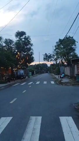 A video captures a street scene at dusk. The camera is positioned at a low angle, suggesting it is attached to a moving object, possibly a bicycle or scooter. The road is paved and has a pedestrian crossing marked with white stripes. On the left side of the road, trees line the path, and a few motorcycles are parked. Some string lights are visible, adding a festive touch. On the right side of the road, a sidewalk runs alongside a brick wall. A person is seated at a small table on the sidewalk, seemingly engaged in an activity. Further down the road, a person is walking. The sky is overcast with hues of orange and pink indicating the sunset. Power lines crisscross the sky. The overall impression is of a quiet, possibly rural or suburban, street during twilight.