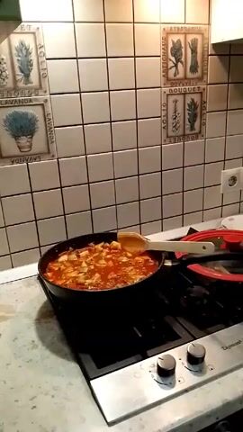 The video shows a black frying pan on a gas stovetop filled with a simmering stew. The stew appears to contain diced zucchini and some type of meat in a red sauce. A wooden spoon rests inside the pan. A red silicone lid is also present on the stovetop next to the pan. The stovetop is stainless steel with black burner grates. The background is a kitchen wall tiled with square white tiles decorated with botanical illustrations and lettering.