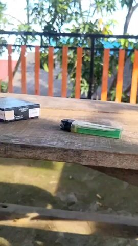Here is a description of the video:

The video shows a wooden table on a porch or balcony with a view of trees, houses, and a blue sky. On the table are a pack of "Magnum" cigarettes, a green lighter, and a glass with some coffee residue at the bottom. The camera zooms out and pans upwards, revealing a railing and a shaded roof above the table. There are no people visible in the video.