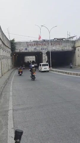 A slightly overcast sky hangs over a concrete underpass. The underpass is marked with "UNDERPASS O BITUNG" in red letters and "SBSN" in blue. Vehicles, mostly motorcycles and a few cars, are traveling through the underpass and on the road leading to it. The road is asphalt, with a white lane marker visible on the left side. Concrete walls line the sides of the road and the entrance to the underpass. Several people are riding on the motorcycles, some wearing helmets, and others are passengers. The scene appears to be a busy urban environment.