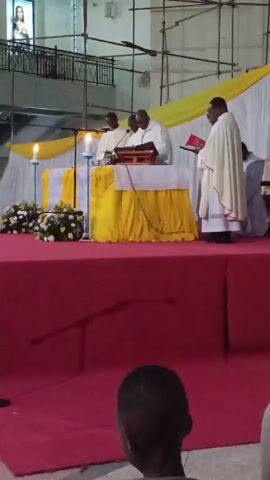 The video shows an indoor religious ceremony. On a red carpeted platform, three priests stand behind a table draped with a yellow cloth. They appear to be conducting a service, one reading from a book on the table while the others listen and read. There are white and yellow floral arrangements on either side of the table, and two candles are lit. A man in priestly garments stands to the right of the table, holding a red book. Behind the platform, white drapes are accented with yellow fabric. In the foreground, the back of a young person's head is visible, suggesting they are part of the audience. The overall environment suggests the inside of a church, with scaffolding visible in the background indicating ongoing construction or maintenance. A statue or painting is visible on a balcony area above.