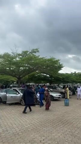 This video shows an outdoor gathering in what appears to be a parking lot. The ground is paved with interlocking bricks, and there are several cars parked. Trees with green foliage line the background. A group of people, some dressed in traditional African attire, are milling about. Some are standing near the cars, while others are in conversation. The sky is overcast with dark clouds, suggesting the possibility of rain. One woman in the foreground is wearing a green and striped dress and jewelry. Another woman in a purple dress is visible interacting with a car. The overall atmosphere seems to be that of a social event or a gathering.