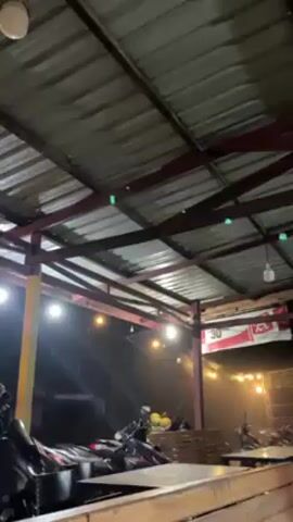 The video depicts an outdoor sheltered area during what appears to be a heavy rain shower at night. The camera is positioned under a sturdy, corrugated metal roof, supported by red-brown painted metal beams. Water droplets are visible on the roof structure, suggesting rain is falling on it.

Beyond the immediate shelter, heavy rain is clearly visible, illuminated by various light sources. There are several warm, yellow-toned bare light bulbs hanging from the ceiling structure, casting a soft glow. In the background, brighter, white floodlight-like sources intensely illuminate the falling rain, creating a visible sheet of water. The sound of the heavy rain is prominent throughout the video.

In the foreground, hints of wooden tables suggest a seating area, possibly for a cafe or food stall. Further back, under the shelter and extending into the rain, several motorcycles are parked. Near the motorcycles, a wooden pallet-like structure is visible, along with a small pile of yellow or orange fruit-like objects. A red and white banner or sign is partially visible in the background, bearing the number "30" and some indistinct text. The overall atmosphere is dim but illuminated by the artificial lights and the reflections off the wet surfaces.