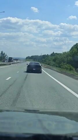 The video shows a dark-colored sports car on a highway, with smoke billowing out from the rear wheels.  The camera is positioned behind the car, presumably inside another vehicle. The car appears to be accelerating or performing a maneuver that is causing tire smoke.  The highway has multiple lanes, with some other vehicles visible in the distance. The environment includes trees along the side of the road and a partly cloudy sky.