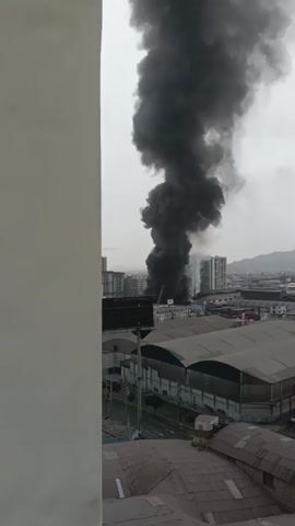 The video shows a large plume of black smoke rising from an urban area, obscuring the sky and the upper portions of buildings. The smoke is thick and billows upwards, indicating a significant fire. In the foreground, the edge of a white structure, possibly a building or wall, takes up the left side of the frame. Behind this, the rooftops of several industrial-style buildings with curved roofs are visible, with some featuring metal siding. Further in the distance, a cityscape with multi-story buildings, some appearing to be residential or office towers, is visible. A fire truck ladder can be seen extended towards the source of the smoke, suggesting emergency services are on the scene. The overall scene is one of an active fire incident. The weather appears overcast, contributing to the muted lighting. The presence of urban infrastructure, including roads and buildings, points to a city environment. There is no visible text in the provided frames. The event is clearly a fire emergency.
