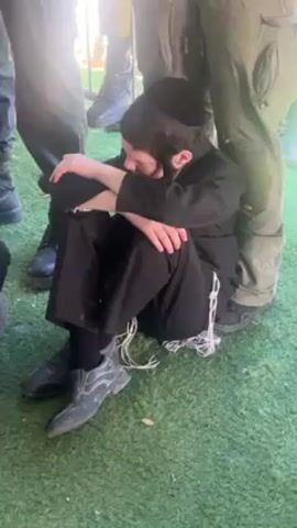 A young boy wearing a yarmulke and tzitzit sits on green artificial grass, surrounded by people, some of whom appear to be in uniform. He is wearing a black suit, black socks, and grey shoes. His hands are crossed over his legs. He looks down. The people around him are wearing dark shoes or boots. One person wears military-style green pants. The camera view is shaky and is mostly focused on the boy. Voices can be heard in the background.