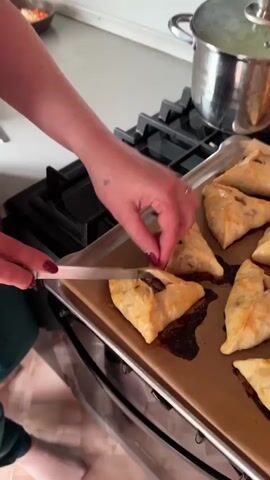 Here's a detailed description of the video:

The video shows a close-up shot of a person's hands using a small knife to cut open a pastry on a baking sheet. The pastries appear to be triangular or square-shaped and golden brown, suggesting they've been baked. Some of them are sitting on burnt drippings on the baking sheet. The filling inside the pastry seems dark, and there's a sprig of green inside. The person has a ring on their finger and red nail polish. A small tattoo can be seen on their hand.

The baking sheet is placed on the stovetop next to a pot with a lid, suggesting a kitchen environment. The stovetop is black and stainless steel. The person's body is only partially visible, and they seem to be wearing a green garment. The pastries are arranged neatly on the baking sheet. The action being performed is carefully cutting open one of the pastries to reveal the filling.