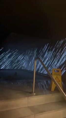The video shows an outdoor scene at night. In the foreground, there are concrete steps leading down. A metal railing runs diagonally across the steps. To the right of the railing, a yellow wet floor sign with a warning symbol is visible. In the background, there is a pool or body of water with blue lights projected onto it, creating a shimmering effect that resembles rain. The water appears to be in motion. The overall environment is dark, illuminated by the blue lights and the sign.