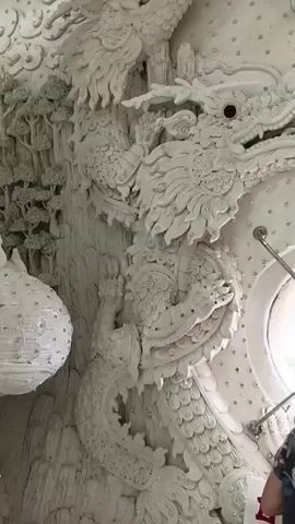 This is a close-up video of a white stucco dragon sculpture on a wall. The dragon has intricate details, with scales, claws, and a ferocious expression. Its eyes are red, and it appears to be coiled and in motion. The background is also adorned with white stucco clouds and other decorative elements. The lighting is bright, highlighting the textures and details of the sculpture.