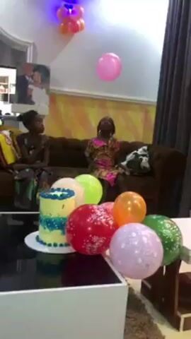 A young girl in a patterned dress is jumping and reaching to play with a pink balloon in a room decorated for a celebration. She is in front of a brown couch where an older woman and another young girl are sitting. On a coffee table in the foreground, there is a decorated cake and a pile of colorful balloons. There is also a set of balloons on the ceiling above the girl. The room has yellow and white patterned walls, dark curtains, and a picture on the wall. The camera is stationary.