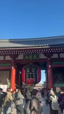The video shows a view of the Kaminarimon Gate at the Sensō-ji Buddhist temple in Asakusa, Tokyo. The gate features a large, red paper lantern with black Japanese characters, and a green sign with Japanese characters is above the lantern. The gate is red and white, with a dark tiled roof. People are walking through the gate into the temple complex. The sky is clear and blue.