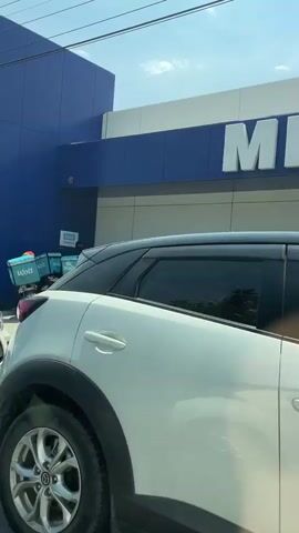 A wide shot captures the exterior of a METRO store. In the foreground, the front of a white car is visible. The car is parked near the entrance, with its side facing the viewer. Beyond the car, a group of people are gathered outside the store. Several motorcycles, many equipped with large blue delivery boxes bearing the Wolt logo, are parked in front of the METRO store. The individuals appear to be interacting with each other, possibly as delivery personnel waiting for orders. The sky above is clear and bright, suggesting a sunny day. The overall scene suggests a busy commercial area with a focus on food delivery services.