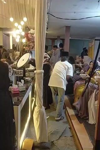 The video depicts an indoor gathering, likely a celebration or event, with several people present. In the foreground, a white drape hangs from the ceiling, partially obscuring a buffet table. The buffet table is set up with various food items, and people are seen serving themselves. To the right, a group of people are seated, some of whom are eating. The environment appears to be decorated, with lights and floral arrangements visible. The overall atmosphere seems to be one of a social gathering where food is being served.