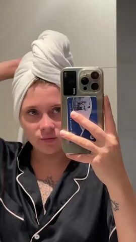 A young woman with blonde hair wrapped in a white towel is taking a selfie in a bathroom mirror. She is wearing dark pajamas with white piping. She is looking at the camera and making faces. She has tattoos on her chest and arm. Her phone is held in her right hand and has a blue and white card attached to the back of the case.