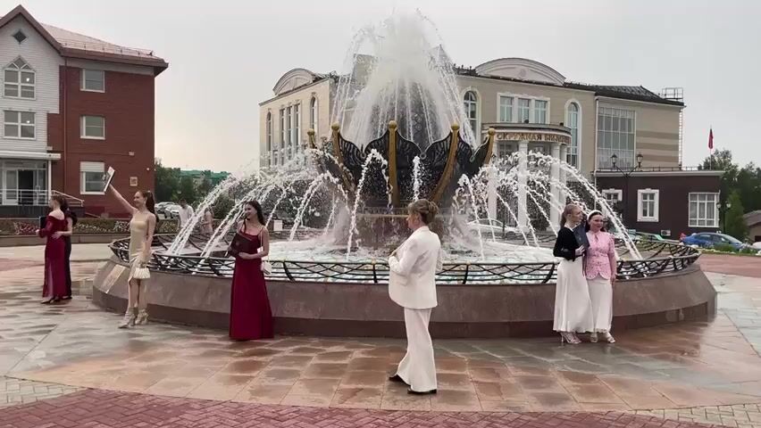 A group of young women in formal attire are gathered around a large, ornate fountain. The fountain features a central sculpture resembling a flower with golden petals, from which water sprays upwards. Several women are standing on the paved area around the fountain. One woman in a red dress holds what appears to be a diploma or certificate. Another woman in a light-colored dress stands to her left, also holding a similar item. In the center, a person in a white suit stands facing away from the camera. To the right of the fountain, two women in light-colored dresses and one in a pink outfit are standing together. In the background, there are buildings with modern architecture, and the sky appears overcast. A man in a blue shirt is visible on the far left, appearing to take a photo. The ground around the fountain is wet, suggesting recent rain. The scene appears to be a graduation or formal event.