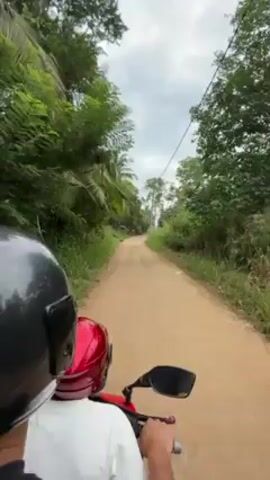 The video shows a first-person perspective from the back of a red scooter traveling on a dirt road. Two people are visible, both wearing helmets: the driver has a black helmet and the passenger in the middle wears a red helmet. The road is surrounded by lush green vegetation, including trees and tall grasses. Palm trees are interspersed among the other trees. Utility poles and wires run alongside the road. A chicken is briefly visible on the side of the road. The sky is overcast with a mix of white and gray clouds. The overall scene suggests a rural or tropical environment.