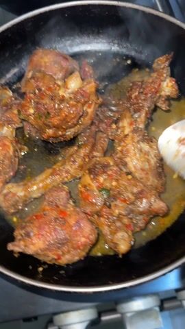 A close-up shot shows a dark-colored skillet on a stovetop. The skillet contains pieces of cooked chicken, coated in a red, spicy-looking sauce. There appears to be some liquid or oil at the bottom of the pan. Steam rises from the chicken. A person is using tongs to turn and move the chicken pieces around in the skillet. The stovetop underneath the skillet is metallic. Another pan is partially visible in the background.