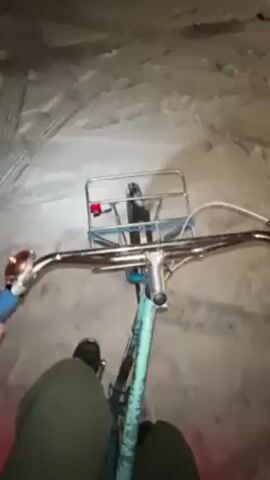 A first-person view shows a person riding a blue and white bicycle on a concrete surface. The bicycle has a metal basket attached to the front. The rider wears dark shoes and green pants. A red light is visible near the back wheel. The rider pedals the bicycle, and the camera angle shifts as they move. The background appears to be a dimly lit, perhaps indoor, space with concrete flooring.