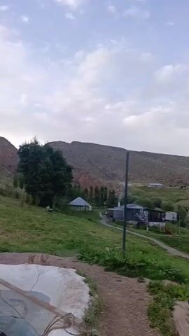 This video shows a clear, shallow pond filled with many small fish swimming. The pond is surrounded by a white, concrete-like border. The pond is located outdoors, with a grassy hill and mountains in the background. The sky is cloudy and the sun appears to be setting. There is a small, covered structure on the hill in the distance.