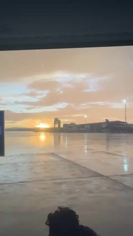 The scene is viewed through a window overlooking an airport runway at what appears to be either sunset or sunrise. The sky is a mix of orange and grey clouds, reflecting on the wet pavement of the runway. A control tower and several airport buildings are visible in the background, along with airplanes. The video appears to be taken from inside a building, and the top of a person's head with curly hair is visible at the bottom of the frame. There are some signs posted on the window, one of which is partially visible. The lighting is somewhat muted due to the cloudy sky and wet surfaces.
