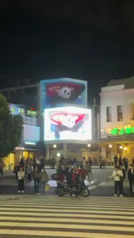 The video shows a busy city intersection at night, with pedestrians, scooters, and buildings clearly visible. The focus is on a pair of large, stacked video screens displaying a 3D animated panda bear. The panda appears to be interacting with the screen, as if reaching out.  Buildings and businesses, including a Starbucks, are visible in the background.  Many people are waiting at a crosswalk.  Traffic moves across the screen.