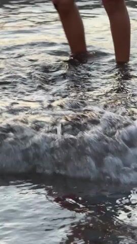 The video shows a close-up, low-angle shot of a person's legs walking through shallow water. The water is clear and reflects the light from above. The person is wearing dark swim trunks with a blue stripe. As the person walks, their feet splash through the water, creating small waves and ripples. The overall impression is one of a peaceful, leisurely stroll along the water's edge.