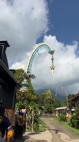 The video shows a large, ornate decorative structure, likely a Penjor, a Balinese offering pole, set against a bright blue sky with scattered clouds. The Penjor is made of bamboo and adorned with white and blue feathers, as well as intricate woven decorations. It arches over a paved pathway and appears to be situated near a building with a dark-colored roof and white walls, which could be a temple or a traditional Balinese compound. Lush green trees and vegetation surround the area, and in the distance, a mountainous landscape is visible. The camera pans upwards, showcasing the full height and design of the Penjor. There are no people clearly visible in the frame, and no specific actions or interactions are depicted. The environment appears to be outdoors, under sunny weather conditions.