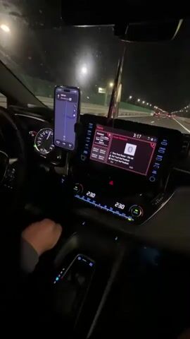 The video shows the interior of a car at night, with the driver's hands visible on the steering wheel and gear shift. A smartphone mounted on the dashboard displays a navigation app. The car's infotainment system shows music playing, with the time displayed as 3:17. The car appears to be on a highway, with streetlights and other vehicles visible outside. The dashboard lights and climate control settings are also illuminated. The overall atmosphere is one of a nighttime drive.