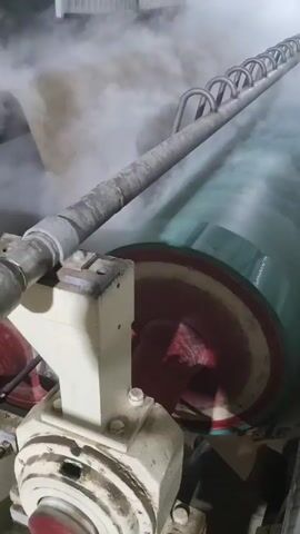 The video shows an industrial setting with a large, rotating drum partially visible, appearing to be part of a manufacturing machine. The drum is green with a red and white striped edge. White steam or vapor is heavily present, obscuring parts of the machine and the surrounding area. The machine also includes metal piping and support structures, painted in light colors. The floor is metal and appears wet. The general atmosphere is one of industrial process, possibly involving heat or steam treatment of materials.