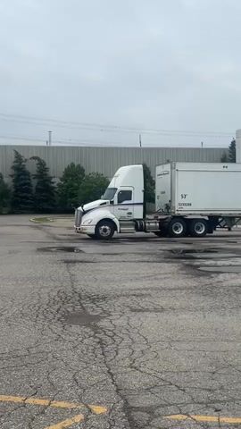 A white semi-truck with a white trailer is parked in an asphalt lot. The truck has a blue stripe on its side. The trailer has the number 53 on its side, as well as some other numbers. There are some trees and a building in the background. The sky is overcast.