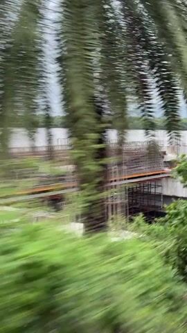 The video shows a construction site next to a body of water. The construction appears to be for a bridge or overpass, with concrete pillars and beams visible. There is also rebar and scaffolding. The water is brown and appears to be choppy. Trees and hills are visible in the background. The video is shaky, suggesting it was filmed from a moving vehicle.