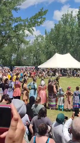 A group of Indigenous women in traditional regalia are gathered in a circle facing a large crowd. They are adorned in brightly colored dresses, beads, and headwear, some holding fans or other ceremonial items. In the background, a large tent provides shade for a seated audience. The event appears to be a Pow Wow, with drums and chanting audible. The sky is blue with scattered clouds, and trees surround the outdoor venue. The camera is held by a spectator in the foreground, capturing the scene from a low angle.