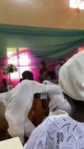 A group of people are gathered indoors, likely for a celebration or event. In the foreground, a woman wearing a white headwrap and a white lace top is partially visible, facing away from the camera. To her left, a man in a white robe is dancing or moving energetically, facing a woman in a white dress. The woman in white appears to be smiling and interacting with the man. Behind them, other individuals are visible. One man in a brown shirt stands with his arms crossed, and further back, a woman in a red dress and yellow headscarf is also present. The environment is decorated with teal and pink drapery hanging from the ceiling, and there are windows with light filtering through. A floral arrangement is visible on a table to the left. The overall atmosphere suggests a lively and joyous occasion.