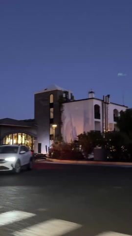 The camera moves past a white car in front of a building at twilight. The building has the words "RIO BACON" in neon lights on the roof. A stop sign is on a post near the car. The camera passes palm trees and shrubs as it drives by the building.