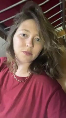 A woman is standing and recording herself in a close-up selfie. She is wearing a burgundy t-shirt and a gold chain necklace. Her brown hair is parted to the side and hangs loosely around her face. She has fair skin with freckles. She looks directly at the camera and touches her hair with her right hand. The background appears to be the underside of a bunk bed frame.