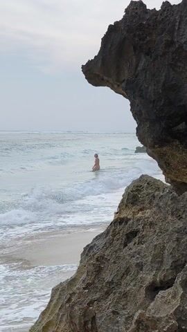The scene is a beach, likely coastal. A person stands in the water with waves gently lapping around them. The person's gender is not clear. The water is a light, clear turquoise, and the sand is white. To the right, a large, dark, jagged rock formation frames part of the view. The sky is overcast. The waves appear small and the person is stationary in the shallow water.