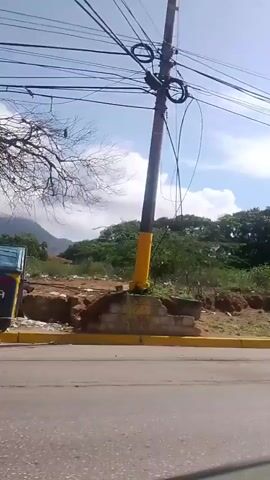 The video shows a roadside view with a clear sky and scattered clouds. The road is paved, with a yellow curb marking the edge. To the left of the road, there is a dry, dusty embankment with sparse vegetation and some debris. Further back, there is a dense line of trees, including palm trees.

As the video progresses, several objects and structures come into view. Initially, there appears to be some discarded items on the embankment, and a handcart with a large wheel is visible. Later, a small, dilapidated building with a corrugated roof comes into view, and a motorcycle is parked in front of it. Further along, a concrete utility pole with numerous wires and cables is prominent. Adjacent to the pole, there is an entrance with a sign that is difficult to read. Finally, the video shows another building with a metal gate and more vegetation. The scene appears to be in a rural or semi-rural area, possibly in a tropical region, given the presence of palm trees and the general appearance of the vegetation and buildings. There are no people visible in the video. The camera is in motion, suggesting it is mounted on a moving vehicle.