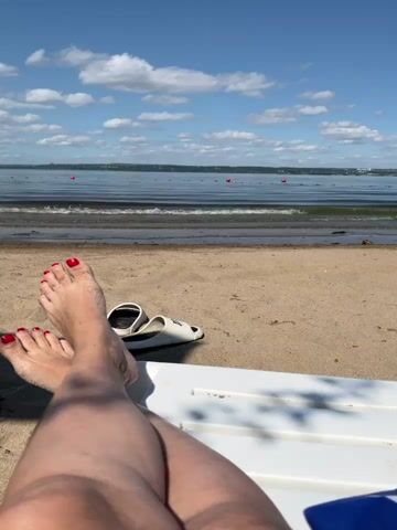 The video shows a close-up of a person's legs and feet relaxing on a beach chair. The person's toenails are painted red. White sandals are placed on the sand next to the feet. The background features a sandy beach, the ocean with gentle waves, and a blue sky with scattered white clouds. Red buoys are visible in the water. The scene appears to be a serene and sunny day at the beach.