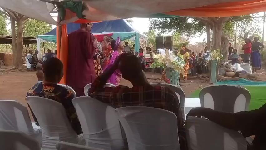 The video depicts an outdoor event, possibly a wedding, under a makeshift tent structure adorned with colorful fabrics. People are seated in white chairs, facing a group of individuals gathered near the front. The attire of those gathered suggests a celebratory occasion. Some people are standing around the seated guests and in the background, watching. Floral decorations and sound equipment are also visible. The environment seems to be a rural area with trees and open space. The camera angle is from behind the seated guests.