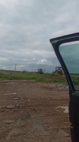 The video shows a field of tall green grass and weeds with three tractors parked in it, shot from the perspective of someone standing near an open car door. The tractors are various shades of blue and black, and all have rotating orange lights on their roofs. Each tractor has a piece of farm equipment attached to its side. The ground in the foreground is dirt with some rocks and scattered garbage. The sky is overcast with white clouds. There are also some power lines visible in the distance.