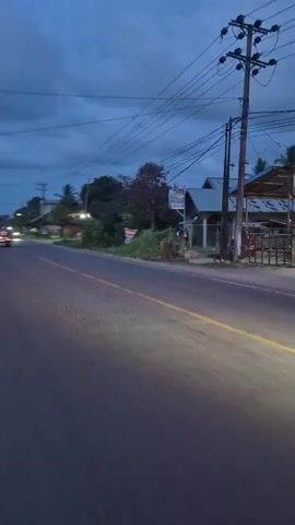 The video shows an ambulance driving down a street at dusk. The ambulance is white with red lights on top. The sky is cloudy and there are trees and buildings on either side of the street. The road has a yellow line down the center. The ambulance passes by the camera and continues down the road.
