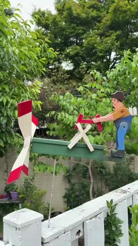 The video shows a wooden whirligig attached to a white picket fence. The whirligig has a green base with a wooden figure of a man chopping wood. The man is wearing a brown hat, a tan shirt, and blue pants. He is holding a red axe and appears to be chopping wood on a wooden X-shaped block. There is a white windmill with red tips attached to the left side of the base, which spins in the wind causing the man to chop. The background includes green trees and plants.